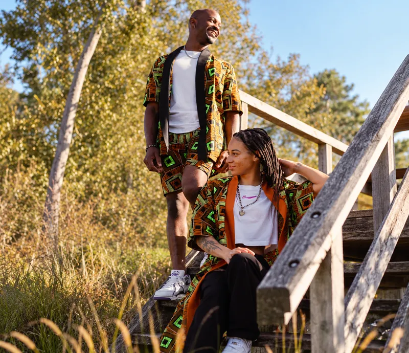 Two customers in matching orange and green print sets on wooden stairs in autumn parkland