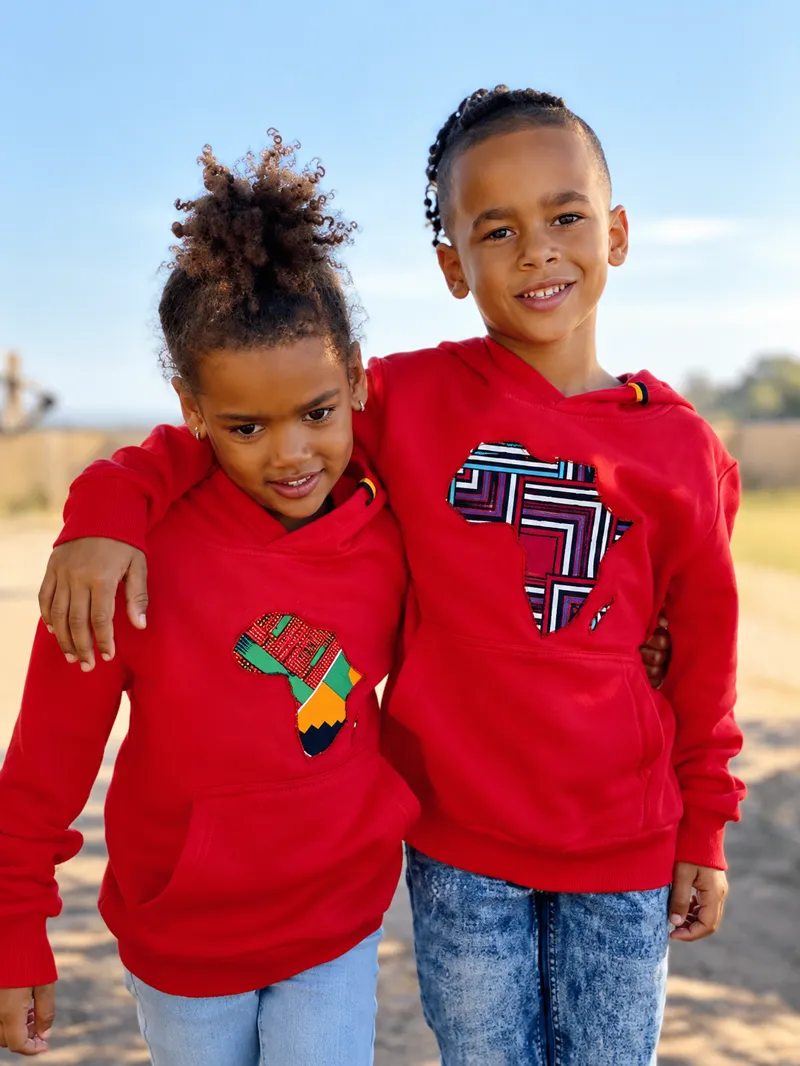 Two children in matching red hoodies with printed Africa motifs, arms around each other on a country road